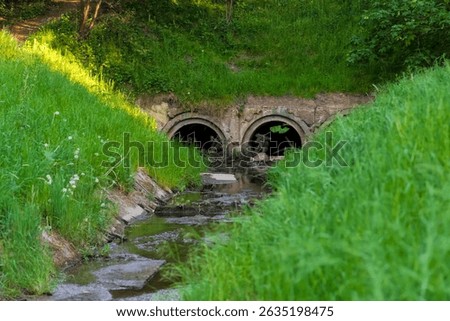 A stream (a tiny river) flowing through a concrete culvert under an embankment slope at a spring sunset. AThe lush, green vegetation runs along the sloped banks under a soft spring sunset.