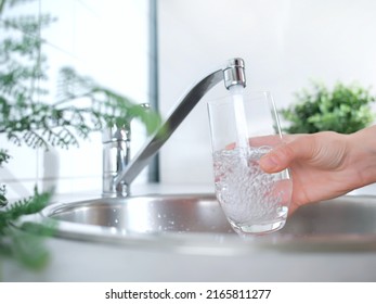 A stream of clean water drink flows into the glass. Woman holding a glass of water under running water from the tap in the kitchen. - Shutterstock ID 2165811277
