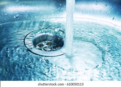 A stream of clean water flows into the stainless steel sink in blue tones. Sink plug hole close up macro.