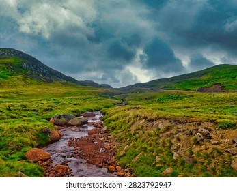 A stream flows gently through a landscape of green hills and rocky terrain. Dark clouds cover the sky, adding depth to the scene during daylight.
