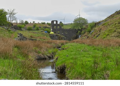 A stream flows through a grassy valley towards the derelict Cheesden Lumb Mill ruins, with wind turbines visible on the distant hills under an overcast sky.