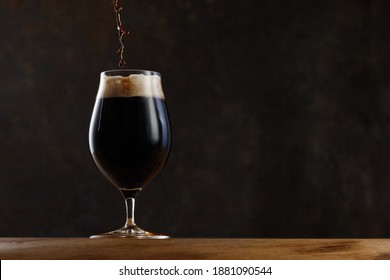 Stream pouring dark beer from the bottle into a beer glass. Jet of the dark porter pours and whips the thick foam in a pint glass against the background of a blurred brown granite wall on wooden table