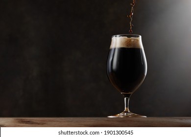 Stream pouring dark beer into a beer glass. Jet of the dark porter pours and whips the thick foam in a pint glass against the background of a blurred brown wall on wooden table - Powered by Shutterstock