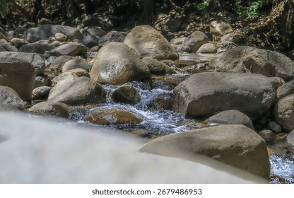 A stream of water flows between large rocks. The water is clear and the rocks are grey. The scene is peaceful and serene