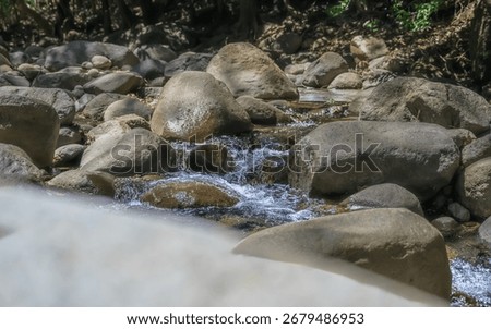 A stream of water flows between large rocks. The water is clear and the rocks are grey. The scene is peaceful and serene