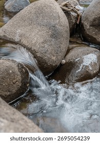 A stream of water flows between two large rocks. The water is clear and calm, and the rocks are grey and rough. The scene is peaceful and serene