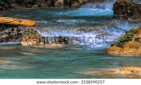 A stream of water flows over rocks and pebbles. The water is clear and blue, and the rocks are brown and gray. The scene is peaceful and serene