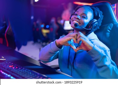 Streamer African beautiful girl shows heart sign with hands professional gamer playing online games computer, neon color.