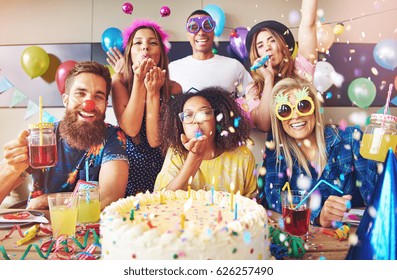 Streamers surrounding group of cheerful friends celebrating a party with large cake and drinks on table in foreground