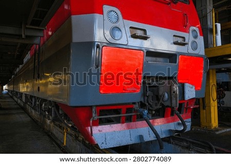 The streamlined train stands on rails in a modern maintenance depot. Its gray and red hull is surrounded by yellow industrial platforms and fences. The locomotive maintenance area.