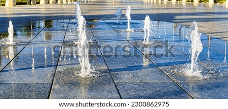 Streams of splashing small fountains on wet paving slabs, illuminated by the sun in a summer park.