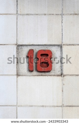 Street address number, number 18. Cracked red wooden number 18 on a dirty white cincerblock wall.