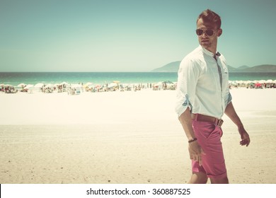 Street Beach Fashion Concept. Portrait Of A Young And Handsome Man In A White Shirt, Pink Shorts Pants And Trendy Glasses Walking Through The White Sand Beach. Close Up. Street Shot.