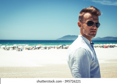 Street beach fashion concept. Portrait of a young and handsome man in a white shirt, pink shorts pants and trendy glasses walking through the white sand beach. Close up. Street shot.