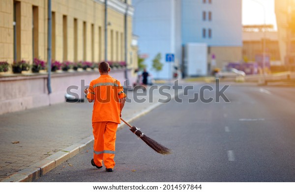 Street cleaner sweeping road and pedestrian zone\
in evening. Municipal worker sweep road with broomstick and\
collects garbage. Sanitation worker clean up street. Utility worker\
cleaning city street