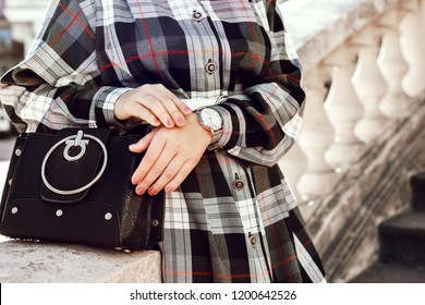 Street close up photo of trendy fashion details of elegant autumn outfit: stylish black bag, silver wrist watch, woman wearing checked dress. Copy, empty space for text