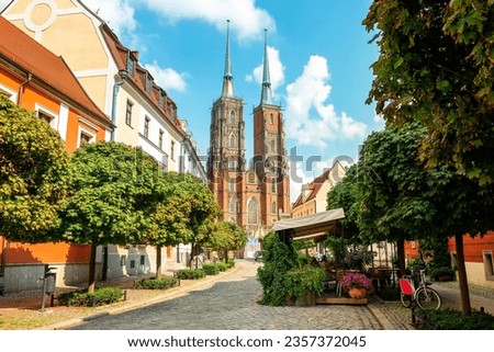 Street with cobblestone road with green trees, colorful buildings, summer cafe, Cathedral of St. John the Baptist church with two spires in old historical city centre, Ostrow Tumski, Wroclaw, Poland