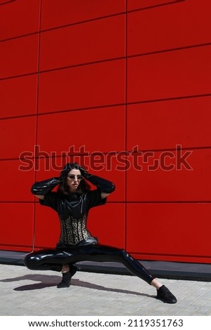 Street fashion concept portrait of beautiful woman with make up. Wearing black latex leather shirt and tights, gloves and corset. Red wall background. Artsy bohemian rock style. no retouch or filter.