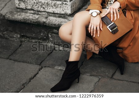 Street fashion details of elegant outfit: woman wearing wrist watch, ankle sock boots, holding light brown, camel color suede handbag, posing in street of european city. Copy, empty space for text