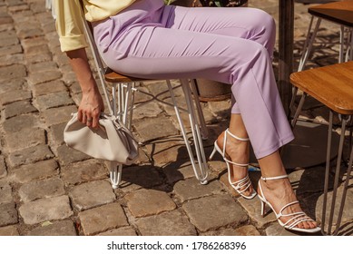 Street Fashion Details Of Elegant Woman`s Summer Outfit: Lilac Trousers, White Strap Sandals, Leather Pouch Handbag