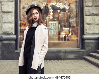 Street Fashion Photo. Beautiful Young Woman In Oversize Coat And Black Hat Over Storefront.