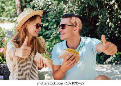 Street Food Concept. Cheerful Happy Couple Holding Hot Dog, Showing A Thumb Up, Sitting Outdoors. Dressed In Summer Clothes, In Sunglasses.