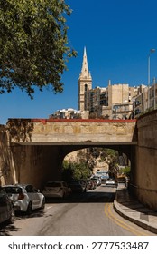 A street level perspective looking through a stone overpass toward the towering spires of Valletta, Malta. Parked cars line the narrow road under a bright sky.