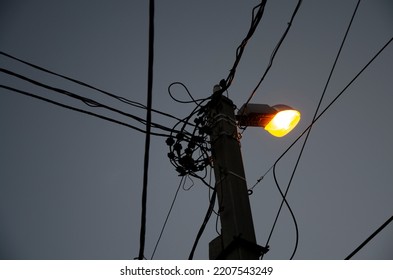 Street Lighting In The Night. A Pole With A Lantern And Randomly Hanging Electric Wires Against Gray Sky. Electrical Cable.