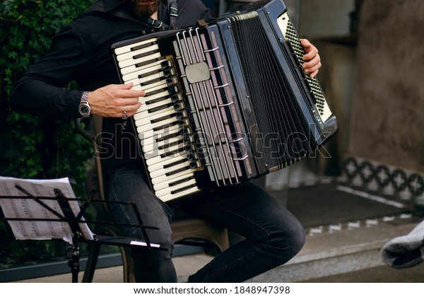 Street musician playing accordion,
stylish man cheers up with beautiful music and earns money
traveling the world, young man plays near an expensive
restaurant