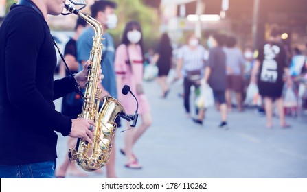 A street musician plays the saxophone and wearing face shield with blurry many people wearing mask and walking in Bangkok, Thailand. The concept of preventing Covid 19 and musical adjustment.