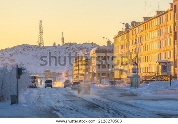 Street in a northern city in the Arctic. Winter\
city landscape. Cars drive along a snow-covered street. Buildings\
at sunset. Telecommunication towers on the mountain. Anadyr,\
Chukotka, Siberia,\
Russia