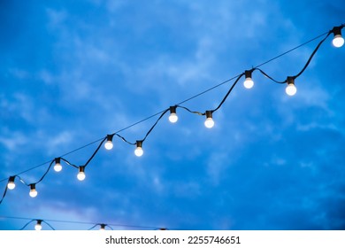 Street outside lighted retro light bulbs garland on night sky. String of light bulb decoration for outdoor activities, party, concert, festival, fun fair. - Shutterstock ID 2255746651