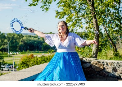 Street performer, beautiful young woman in blue skirt and white blouse, with soap bubbles shows soap show in summer in open air park for children