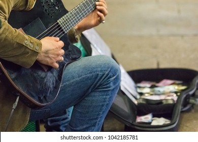 Street Performer Plays Guitar At Night  - Shallow Focus With Blur Background Of His Guitar Case Full With Banknotes And Coins