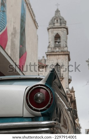 A street photo of a close up on a backlight of an old American car and a bell tower of a church in Havana - Cuba during a rainy day 