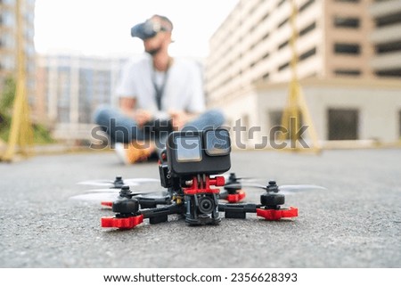 Street photo of fpv multicopter drone landing on the street road with the male pilot operating on the background. Focus is on copter.