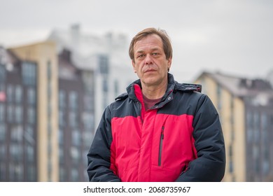 Street Portrait Of A 45-50-year-old Man With A Serious Expression On The Background Of Urban High-rise Buildings, Medium Plan. Maybe He Is A Military Pensioner Or An Actor, A Truck Driver, A Buyer.