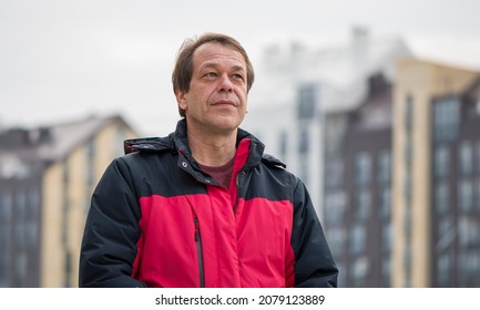 Street Portrait Of A 45-50-year-old Man With A Serious Expression On The Background Of Urban High-rise Buildings, Medium Plan. Maybe He Is A Military Pensioner Or An Actor, A Truck Driver 1.