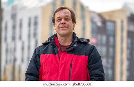 Street Portrait Of A 45-50-year-old Smiling Man Against The Background Of Urban High-rise Buildings, Medium Plan. Maybe He Is A Military Pensioner Or An Actor, A Truck Driver, A Buyer Or A Loader.