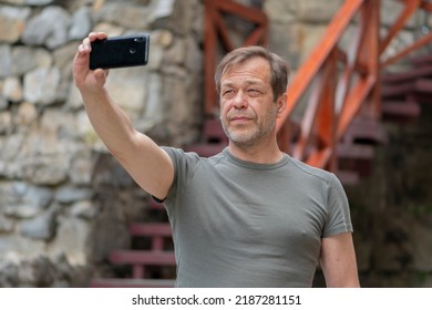 Street Portrait Of An Elderly Man 45-50 Years Old In A T-shirt In Summer, Taking A Selfie On His Phone Against The Background Of An Old Stone Wall.