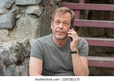 Street Portrait Of An Elderly Man 45-50 Years Old In A T-shirt Talking On A Mobile Phone In The Summer Against The Background Of An Old Stone Wall.