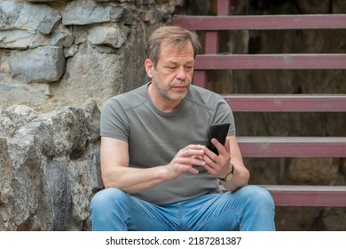 Street Portrait Of An Elderly Man 45-50 Years Old In Summer, Looking Through Messages On A Mobile Phone Against The Background Of An Old Stone Wall.