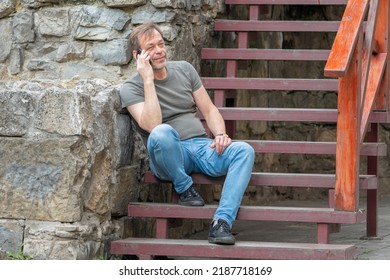 Street Portrait Of An Elderly Man 45-50 Years Old In A T-shirt Talking On A Mobile Phone In The Summer Against The Background Of An Old Stone Wall.