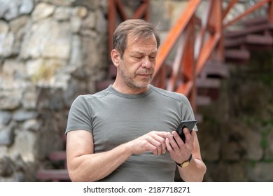 Street Portrait Of An Elderly Man 45-50 Years Old In The Summer, Looking Through Messages On A Mobile Phone, Sitting On The Steps.