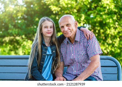 Street Portrait Of Granddad And Granddaughter Sitting On Wooden Bench In City Park At Sunny Summer Day. Grandparent And Grandchild Relationship Concept.