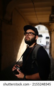 Street portrait of a male photographer with a beard in glasses and a cap with a vintage camera. Young hipster man with digital vintage camera and backpack looks up.
