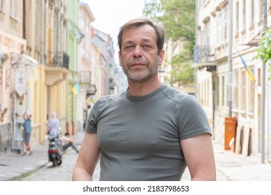 Street Portrait Of A Muscular Man 45-50 Years Old In A Green T-shirt On A Neutral Urban Blurry Background. Perhaps He Is An Elderly Buyer, An Actor Or A Truck Driver, A Loader Or A Military Pensioner.