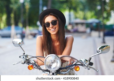 Street portrait of pretty smiling girl on motorbike. Fashion young woman wearing sunglasses and stylish hat on a retro scooter on a summer day in a cityspace.