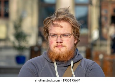 Street Portrait Of A Serious Blond 25-30 Year Old Man With Glasses And A Beard On The Background Of The City, The View Is Directed To The Camera.