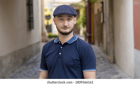 Street Portrait Of A Serious Young Man 30 -35 Years Old In A Cap With A Beard Looking Directly Into The Camera On A Neutral Background Of The Old City,